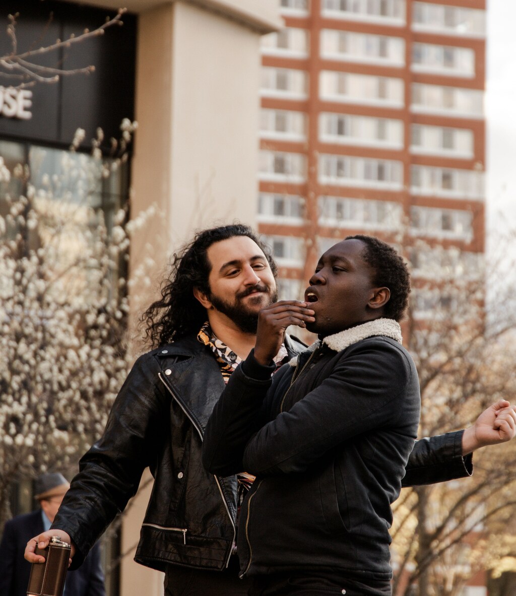 an actor with long black hair and a black beard wearing an orange, pink, and black floral shirt and a black leather jacket and holding a brown leather and metal flask approaches with wide open arms another actor with short black hair wearing a fur collared denim jacket who is stroking his chin in deep thought