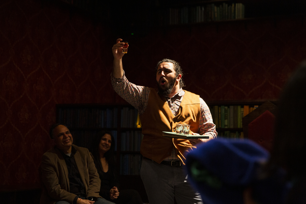 an actor with black hair and a black beard wearing a checked button up shirt, a tan vest, and blue pants holds a tray of shot glasses in one hand and a single shot glass aloft in the other hand as he makes a toast