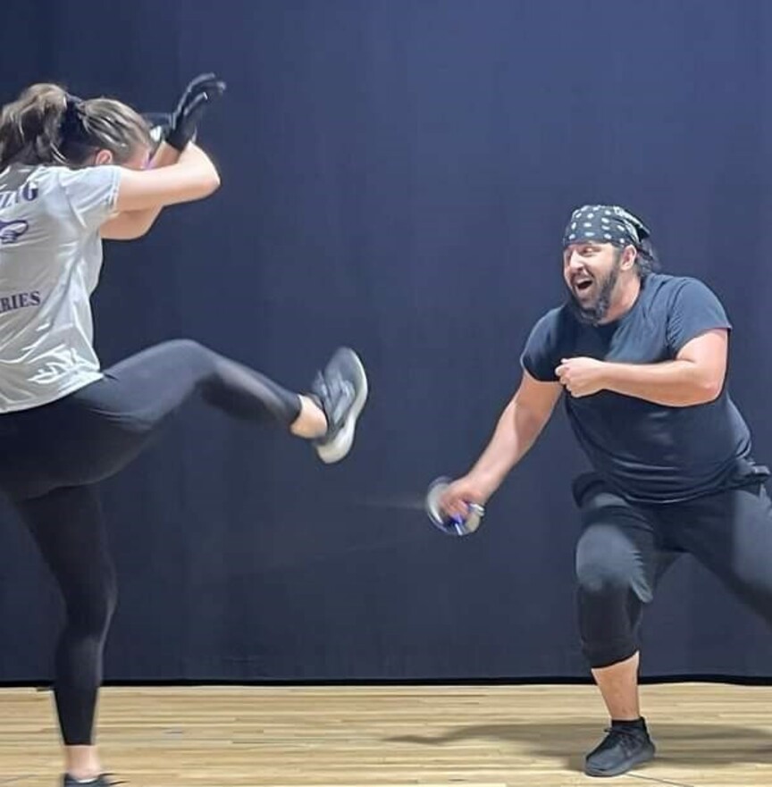 an actor with a black beard and a black bandana on his head wearing a black t shirt, black pants lunges forward and swipes a rapier at the feet of another actor with a brown pony tail wearing a grey shirt, black pants, black shoes, and black gloves who is reeling back on their left leg with their right leg suspended in the air to avoid the attack 